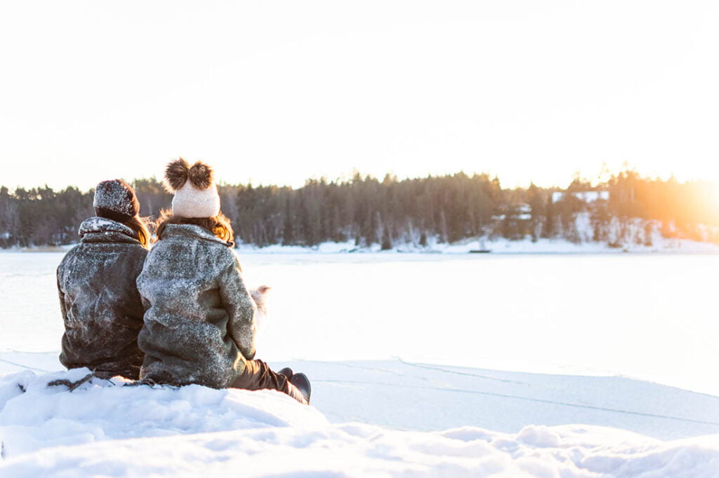 Pariskunta istuu lumen päällä meren edessä katsoen aurinkoon kauniina talviaurinkopäivänä.