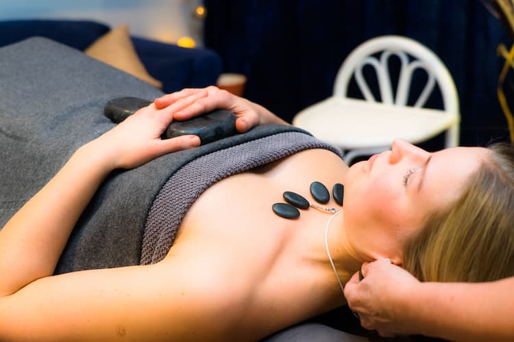 En kvinna ligger på en spasäng och får massage med varma stenar, med stenar i linje längs hennes bröst och axlar, täckta av en handduk. Hon verkar avslappnad med slutna ögon, i ett mjukt upplyst rum