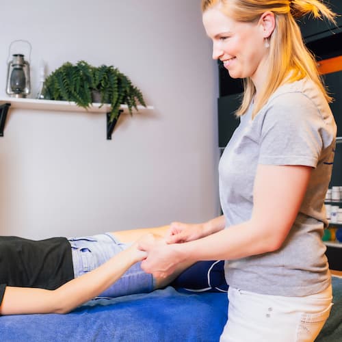 En kvinnlig sjukgymnast med blont hår ler när hon hjälper en patient som ligger på ett behandlingsbord genom att sträcka ut hans högra arm. Rummet har ett modernt utseende med mjuk belysning och gröna växter, förkroppsligande