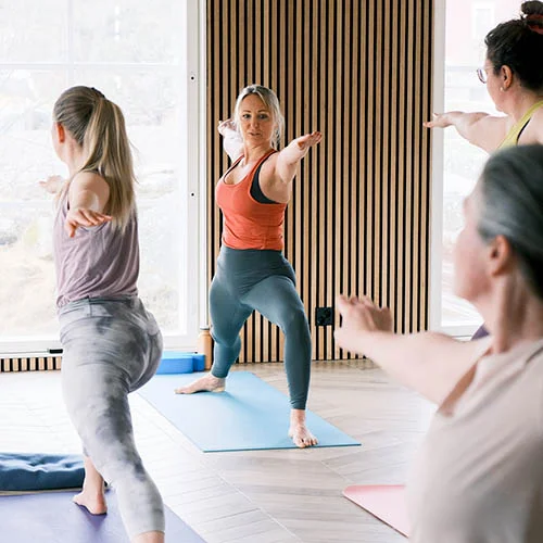 A yoga instructor demonstrating a pose to a small group of women in a brightly lit studio with mirrored walls.