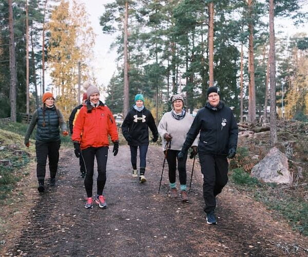 Fem vuxna som går på en skogsstig, klädda i idrottskläder i kallt väder, ler och njuter av att vara utomhus.