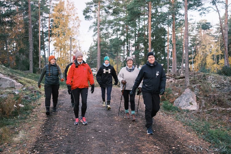 Fem vuxna som går på en skogsstig, klädda i idrottskläder i kallt väder, ler och njuter av att vara utomhus.