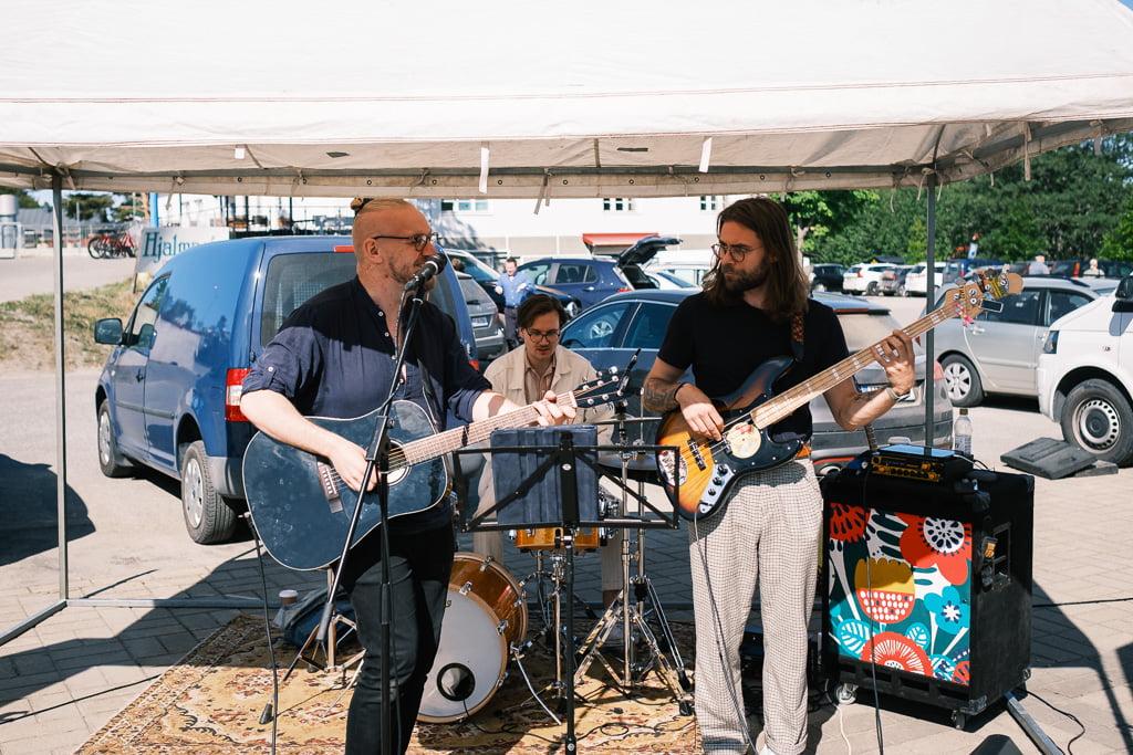 Three musicians perform under a roof in a car park. One plays acoustic guitar and sings, another plays drums and the third plays electric bass. Several vehicles are visible in the background.