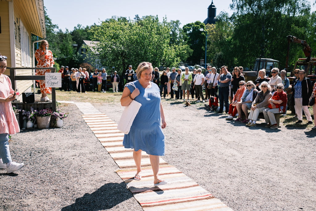 A woman in a light blue dress walks along a makeshift track outside while people sitting and standing nearby watch her.