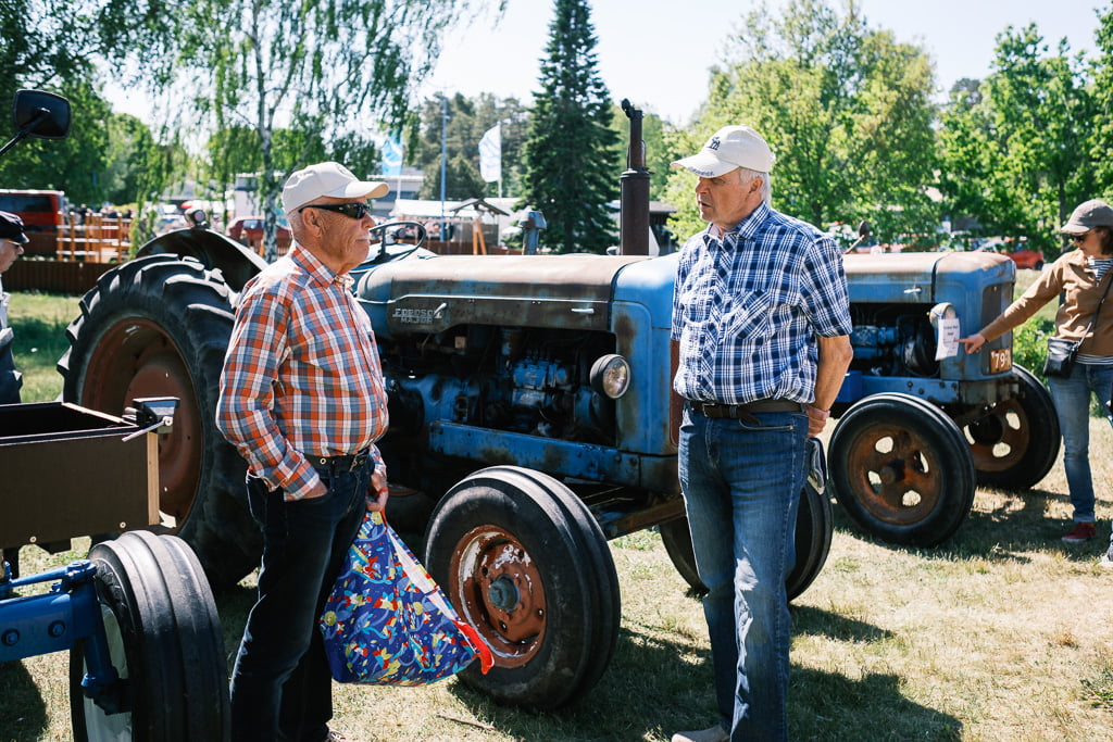 Two men in checked shirts and hats stand talking in front of an old blue tractor at an outdoor event with trees and other tractors in the background.