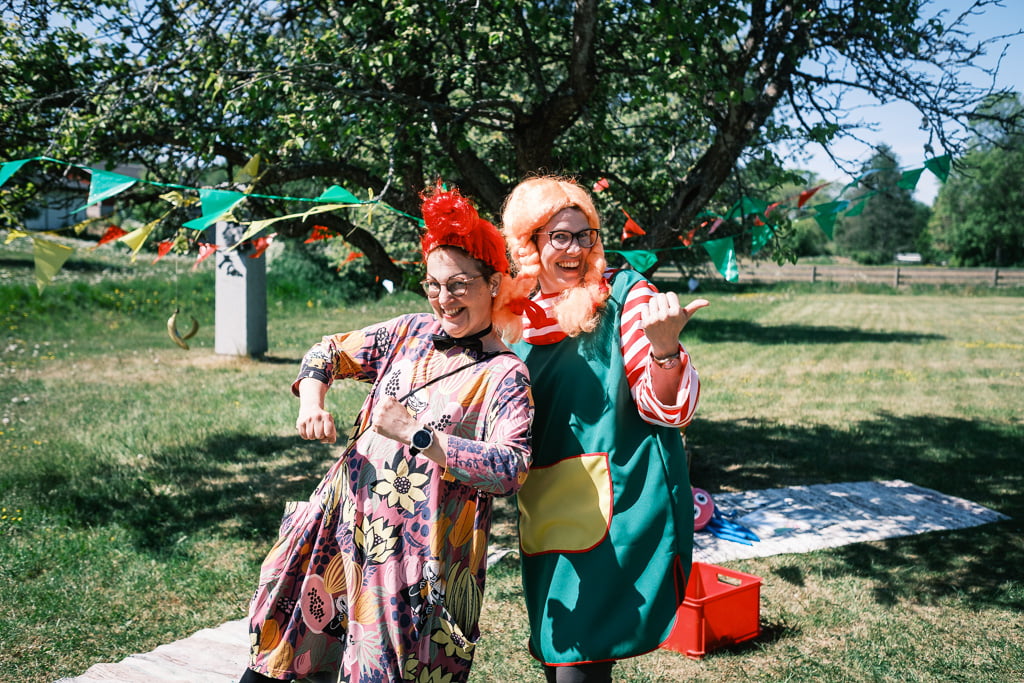Two people dressed in colourful, whimsical costumes pose outside near a tree decorated with vibrant flags. One person gives a thumbs up, and the scene is under a sunny sky.
