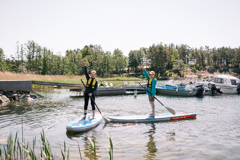 Två personer som bär flytvästar står på paddleboards, höjer varsin arm, på en lugn vattenmassa med flera små båtar anlagda i bakgrunden.