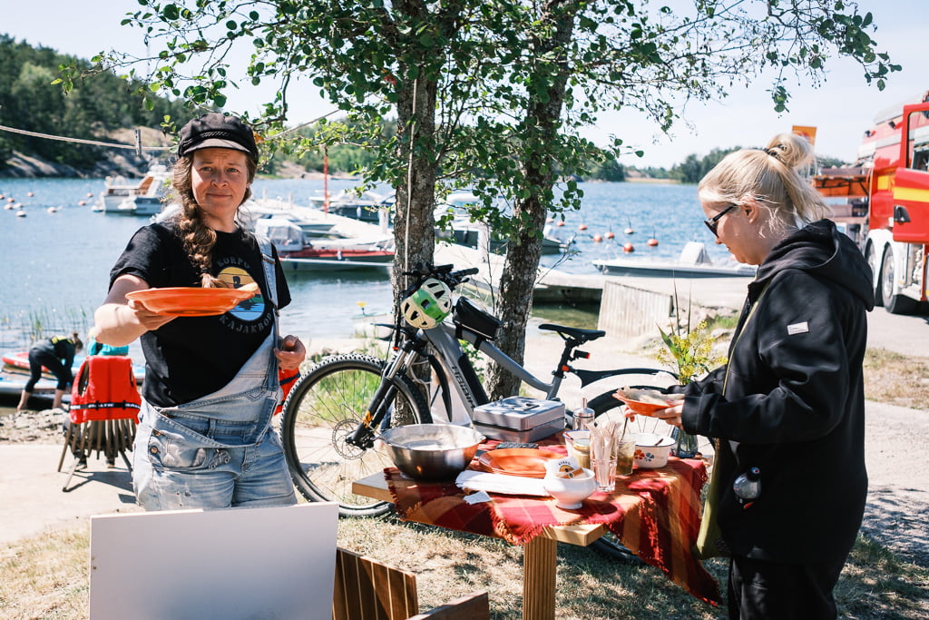 Two people are serving food at an outdoor facility on a beach. The table has various dishes, and a bicycle is parked nearby. Boats are anchored in the background under a sunny sky.