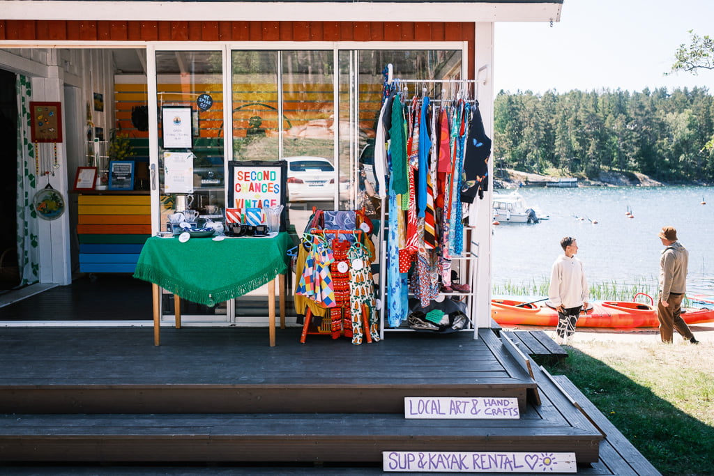En liten butik vid vattnet visar vintagekläder, lokal konst och hantverk. Skyltar visar stand up paddleboard (SUP) och kajakuthyrning. Två personer går vid vattnet med kajaker i bakgrunden.