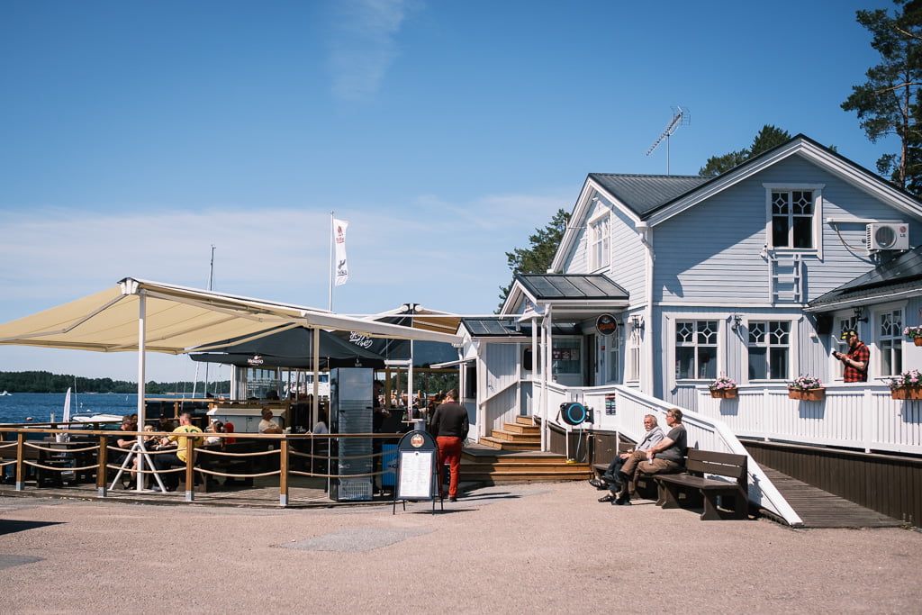 A sunny day in a seaside café with customers eating outside and a white building in the background. Several people are sitting and a person in red trousers is walking towards the café.