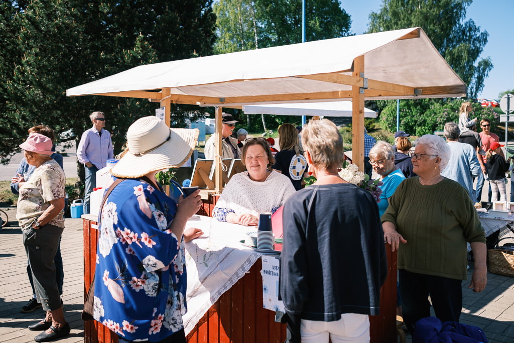 A woman sells goods at an outdoor market stall. People gather around, some interacting with the vendor, on a sunny day with trees in the background.