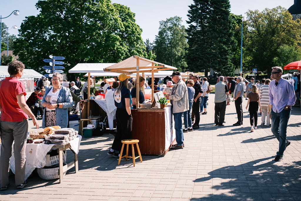 People visiting a sunny outdoor market with various stalls selling food and goods, surrounded by trees and some street signs.