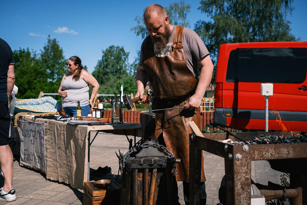 A bearded blacksmith in a leather apron works on an anvil at an outdoor event, with a red van and people in the background.