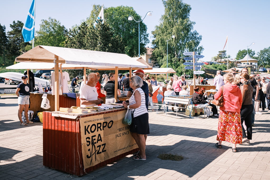Ihmiset katselevat Korpo Sea Jazz -festivaalin ulkoilmamarkkinoiden kojuja, ovat vuorovaikutuksessa ja nauttivat aurinkoisesta päivästä. Markkinoilla on puisia kojuja, joissa on erilaisia tavaroita ja tietoa.