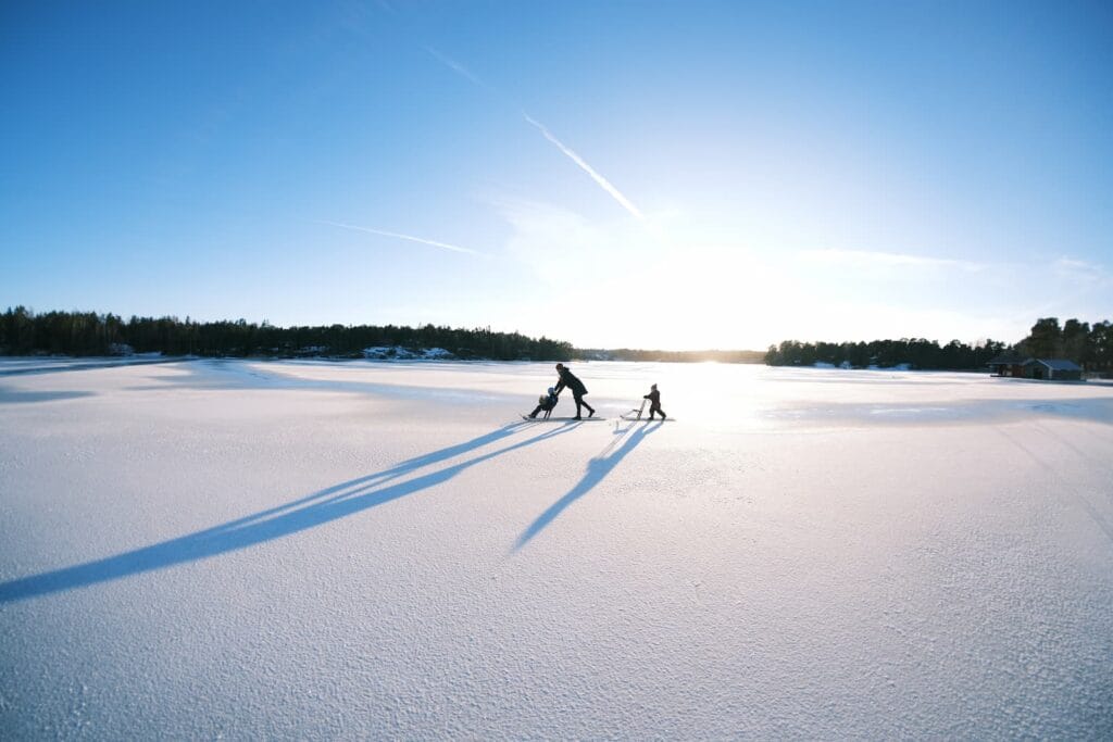 Kaksi ihmistä kelkkailee suurella lumisella pellolla kirkkaalla sinisellä taivaalla, joka heittää pitkiä varjoja auringonvalossa.