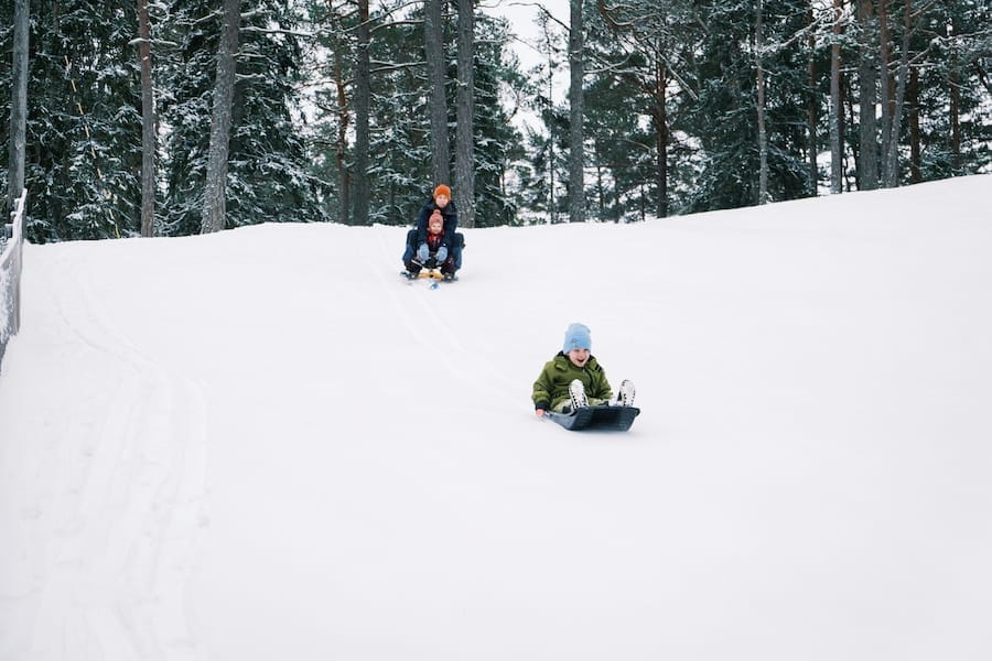 Två personer åker pulka nedför en snöig kulle i ett skogsområde, med snötäckta träd i bakgrunden.