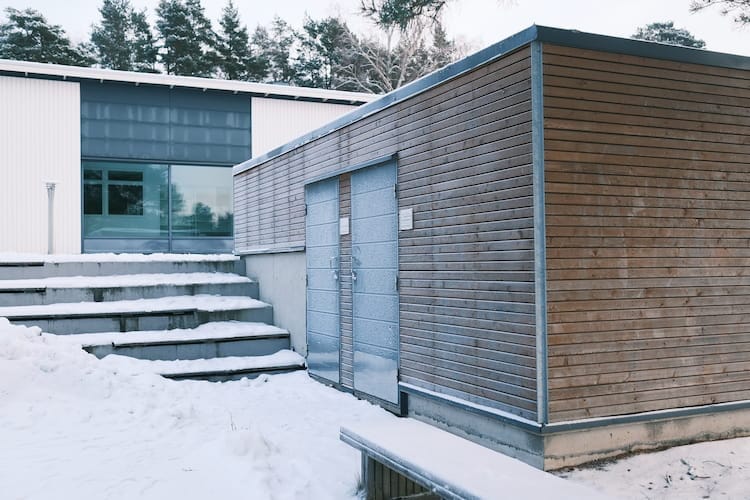 Modern byggnad med träpanelväggar och stora fönster, omgiven av snö. Träden syns i bakgrunden.