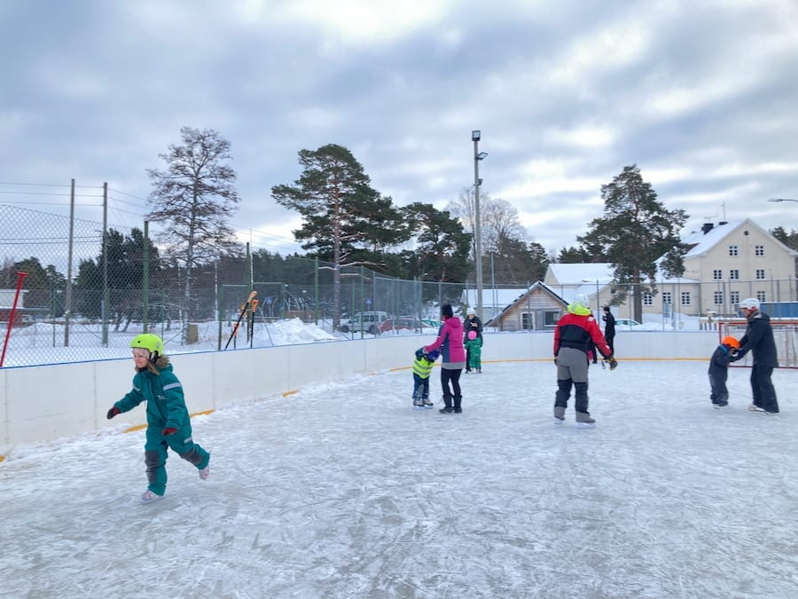 Människor åker skridskor på en utomhusrink omgiven av snö och träd under en molnig himmel.