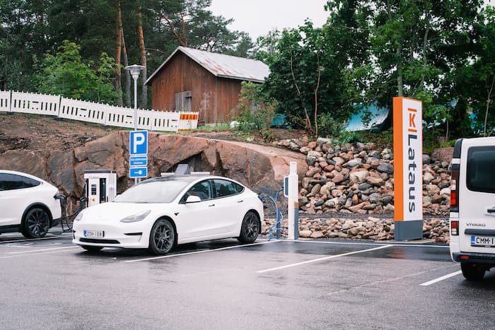 White electric car parked at a charging station on a plot surrounded by trees and a wooden building.