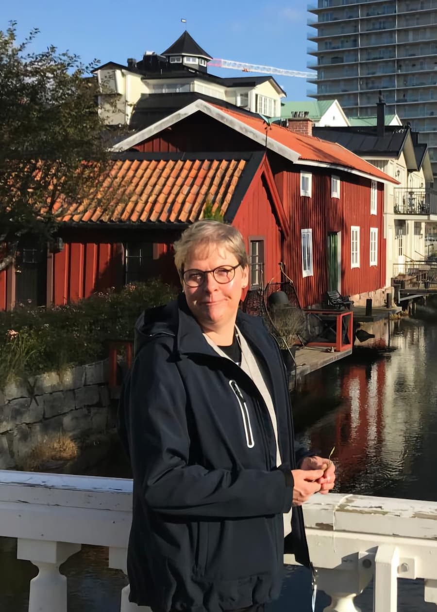 Tina Roswall i mörk jacka som står på en bro framför en röd träbyggnad, med en kanal och andra byggnader i bakgrunden.