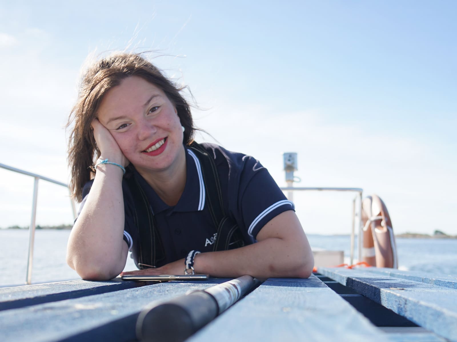 En person med mörkt hår och en marinskjorta ler medan han lutar sig mot en träyta på en båt, med vatten och himmel i bakgrunden.