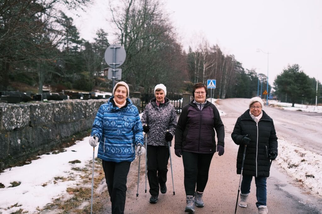 Fyra äldre människor som går med stavar på en snöig stig bredvid en väg.