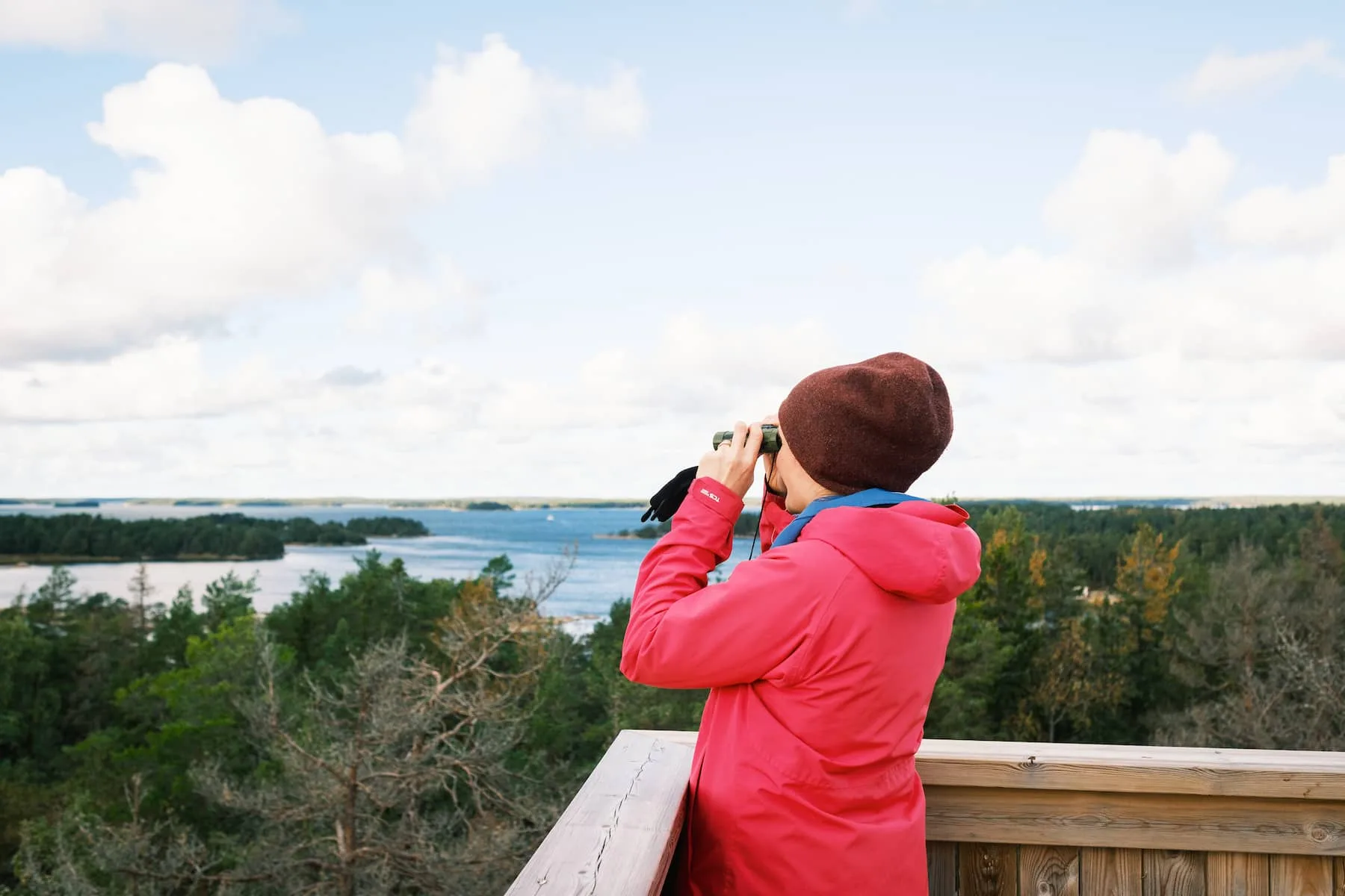 En person i röd jacka och brun hatt står på en träplattform och tittar på landskapet genom en kikare med träd och vatten i bakgrunden.