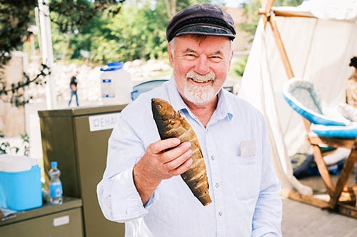En äldre man i keps och ljus skjorta ler medan han håller en rökt fisk utomhus nära tält och campingutrustning.