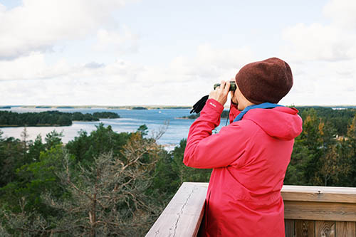 En person i röd jacka och brun hatt står på ett trädäck och använder en kikare för att titta på en sjö och ett skogslandskap.