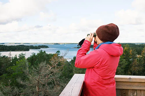 En person i röd jacka och brun hatt står på ett trädäck och använder en kikare för att titta på en sjö och ett skogslandskap.