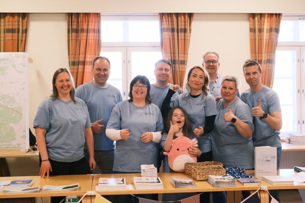 Kahdeksan aikuista, joilla on samanlaiset siniset paidat, ja yksi lapsi poseeraavat ja hymyilevät esitteitä ja pehmoleluja sisältävän pöydän takana kirkkaassa huoneessa, jossa on oranssit verhot.