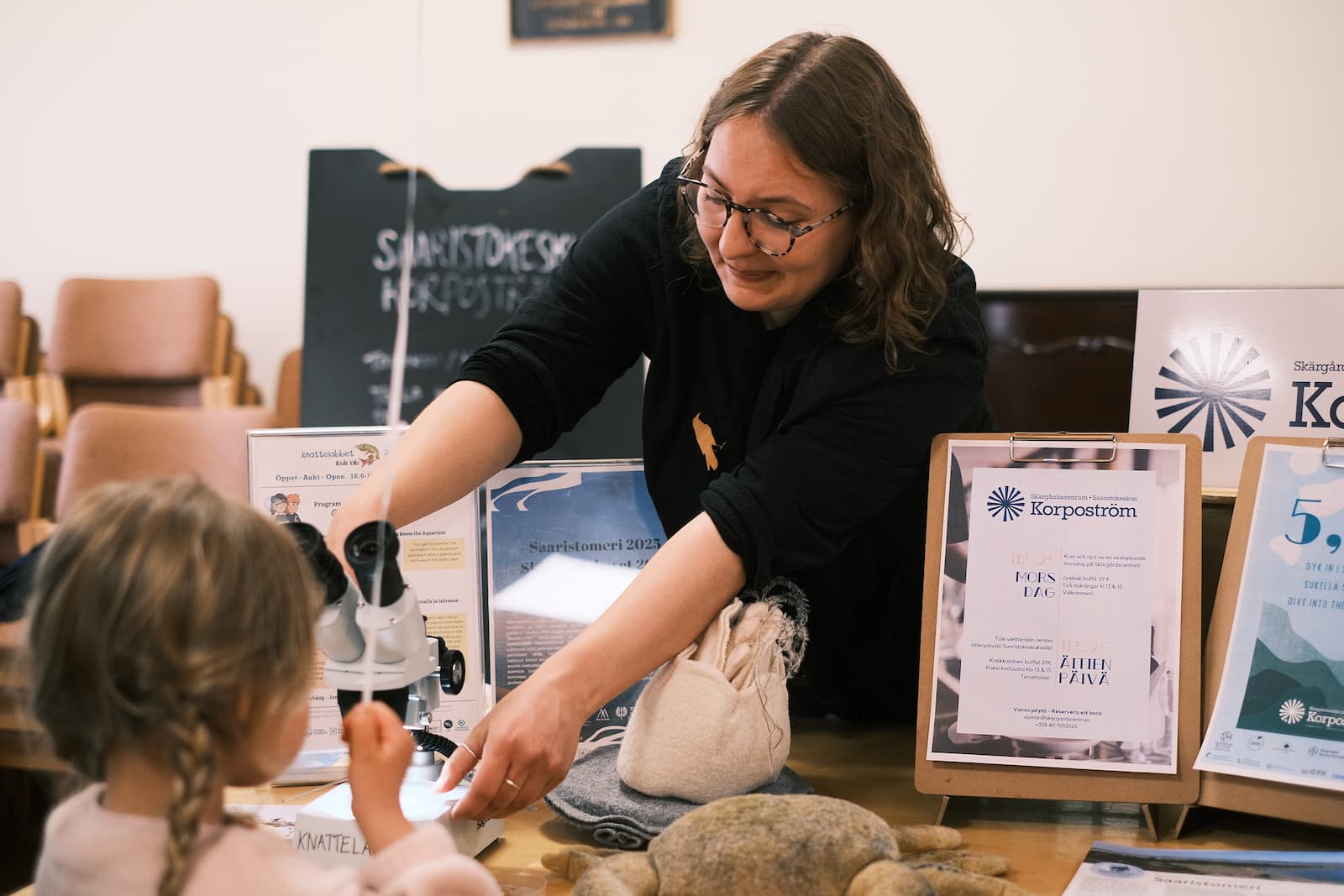 Nainen esittelee pienelle lapselle tiedeaktiviteettia pöydän ääressä, jossa on infonäyttöjä ja opetusmateriaaleja.