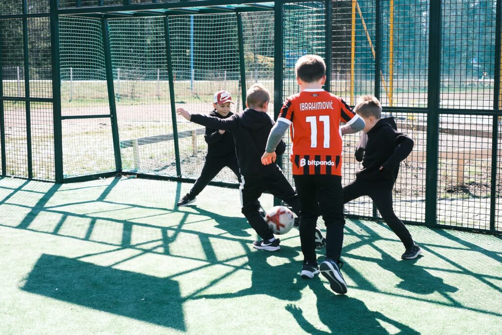 Neljä lasta pelaa jalkapalloa ulkokentällä. Yhdellä lapsella on punainen Ibrahimovic 11 -paita ja hän jahtaa palloa, kun muut yrittävät estää maalin.