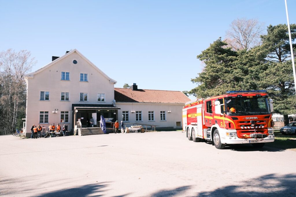 En brandbil står parkerad utanför en ljus byggnad med flera personer i orange västar samlade nära entrén en solig dag.