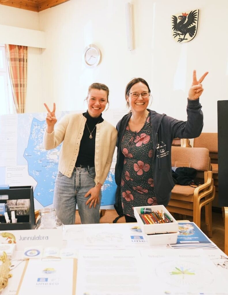 Kaksi naista seisoo näyttelypöydän ääressä, jossa on karttoja ja piirroksia, molemmat hymyilevät ja tekevät rauhanmerkkejä. Pöydällä on taide- ja tiedotusmateriaalia.