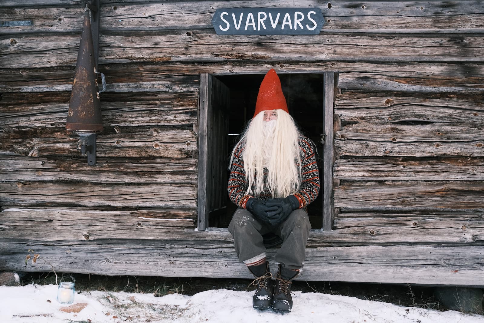 Joulupukiksi pukeutunut henkilö, jolla on punainen hattu ja pitkä valkoinen parta, istuu ikkunassa maalaismainen puumökki, jossa lukee "Svarvars". Lunta ja palava kynttilä ovat maassa ulkona.